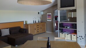 Een moderne woonkamer met een bank, een eettafel met een fles, een televisie, en een keuken met kastjes en een magnetron.