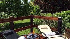 A wooden deck with two lounge chairs, one with a black cushion and one with a white cushion, and a small table with a bottle and a hat. - Petit Vue Vert - One  Bedroom Gite with heated pool on edge of medieval village (Puy-l'Évêque)