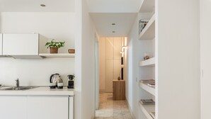 A narrow hallway with a kitchen area on the left, featuring a sink, kettle, and cabinets. A wooden cabinet and a plant are visible on the left wall. The hallway leads to a door at the end. - Torremuzza Home by Wonderful Italy (Palermo)