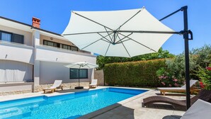A modern outdoor pool area with a large umbrella, lounge chairs, and a well-maintained hedge.