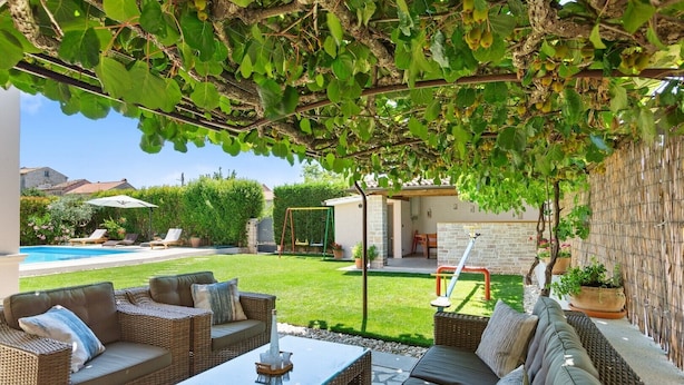 A covered outdoor seating area with wicker furniture and a glass table, overlooking a swimming pool and a garden.
