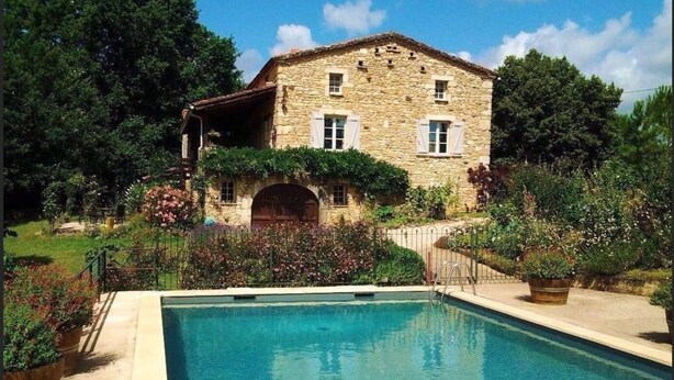 Un edificio de piedra con piscina, rodeado de exuberante follaje y plantas en flor.