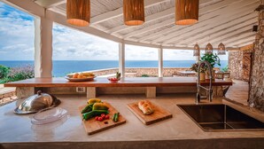 Una cocina con vista al mar, que cuenta con encimera de madera, luces colgantes y una pared de piedra.