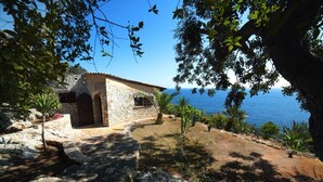 Un edificio de piedra con vistas al mar, rodeado de árboles y terreno rocoso.