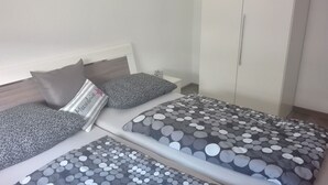 Une chambre avec deux lits, chacun recouvert d’une couette à motifs pois gris et blancs, agrémentée d’un oreiller rayé et d’un oreiller gris. On y trouve une table de chevet blanche avec une lampe et un petit objet décoratif.