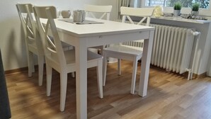 Un coin repas avec une table et des chaises blanches, un radiateur et des plantes en pot sur un rebord de fenêtre.
