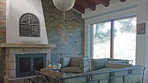 A cozy living room with a stone fireplace, a cushioned sofa, and a large window offering a view of trees.