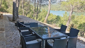 An outdoor dining area with a stone-paved floor, wicker furniture, and a view of a lake surrounded by trees.