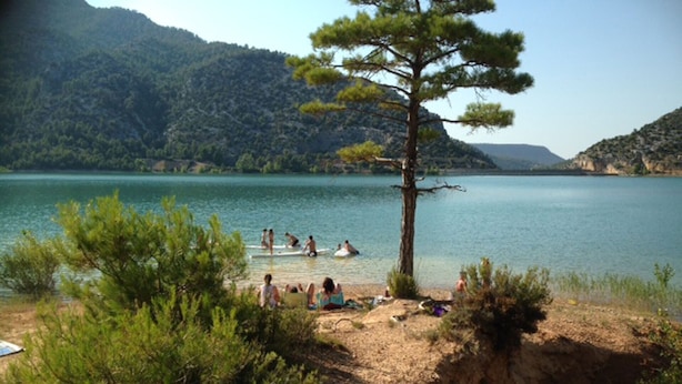 A beach with people swimming and sunbathing, surrounded by greenery and hills.