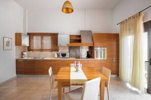 A modern kitchen with wooden cabinets, a dining table, and a large window with curtains. - Blue Wave Villa up to 10 (RHODES)