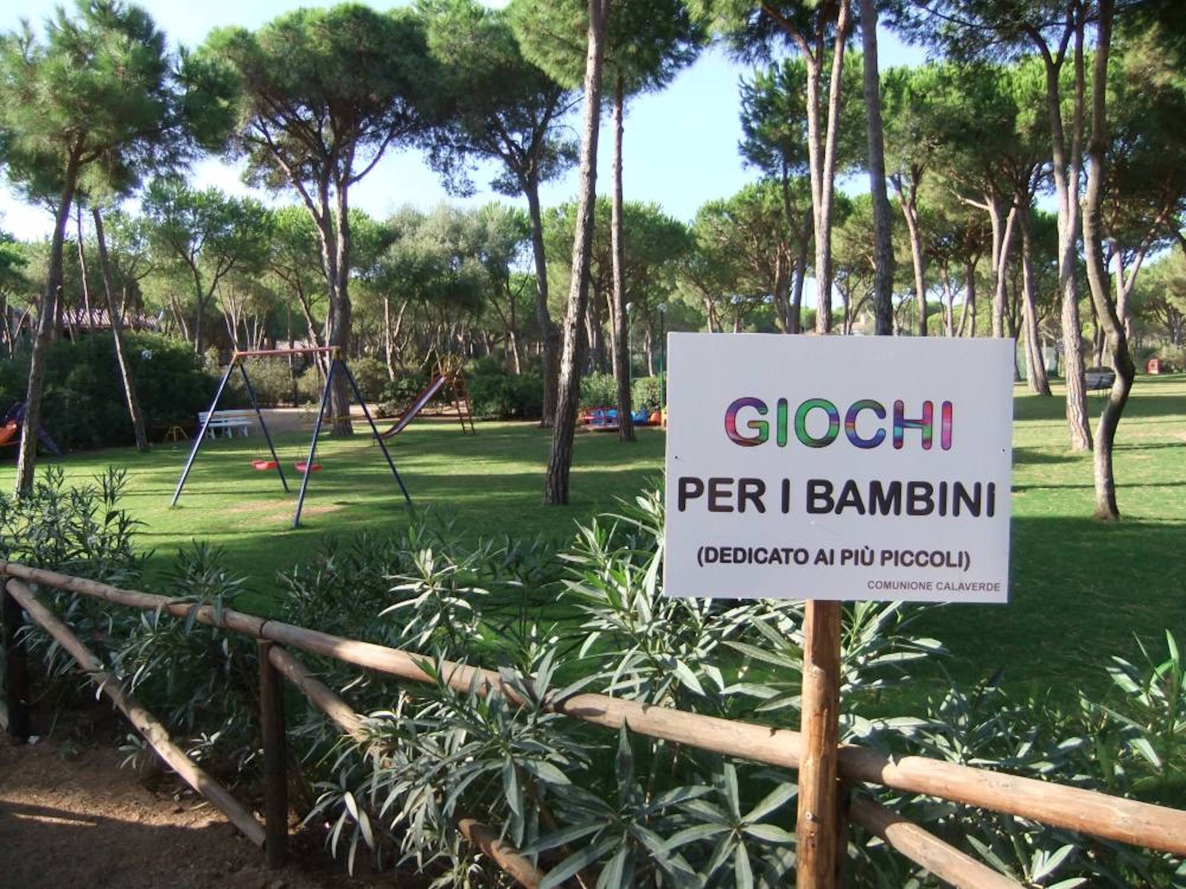 Segnale per un'area giochi per bambini chiamata "GIOCHI PER I BAMBINI" in un parco con alberi e altalene.