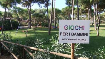 Segnale per un'area giochi per bambini chiamata "GIOCHI PER I BAMBINI" in un parco con alberi e altalene.