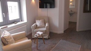 Ein modernes Wohnzimmer mit einer Couch, einem Glastisch, einem flachbildfernseher an der Wand, einem Fenster mit Ausblick und einem Badezimmer mit Waschbecken und Spiegel.