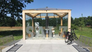 Ein moderner Pavillon mit großen Glasschwingtüren und Holzrahmen, gelegen in einem Park mit Bänken und einem in der Nähe abstellenden Fahrrad.