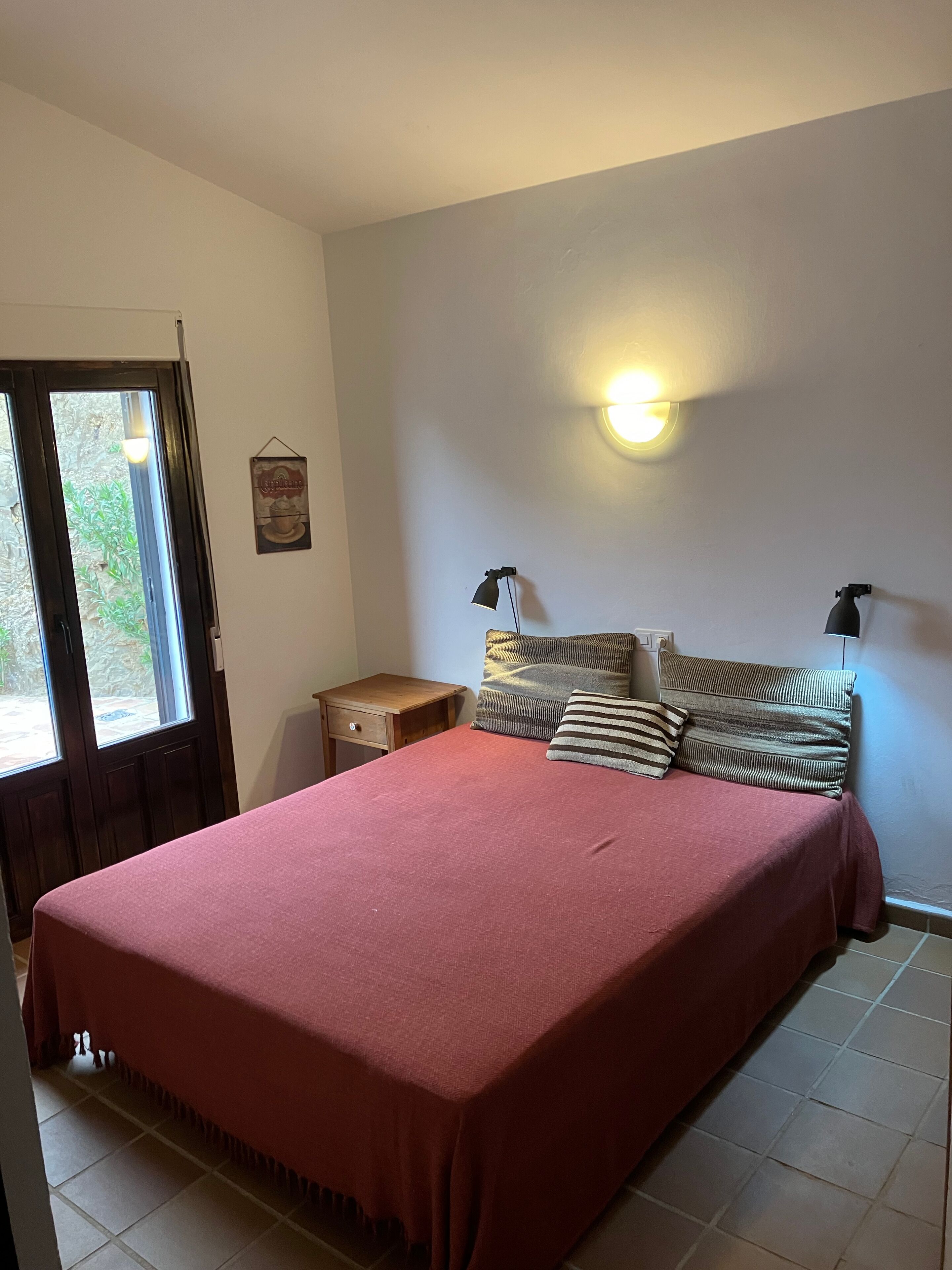Een slaapkamer met een groot bed, een nachtkastje, een wandlamp en een deur die uitkomt op een balkon.