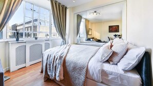Une chambre avec une grande fenêtre, un lit avec du linge de lit blanc, une table de chevet et un miroir.