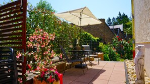 Relax in a serene outdoor space surrounded by lush greenery and flowering plants, with a variety of seating options and a large umbrella providing shade.