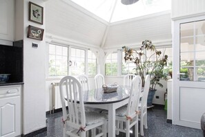 A conservatory with a round glass table and white chairs, surrounded by windows and potted plants.