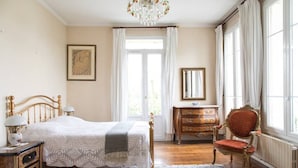 Ein Schlafzimmer mit einem Bett, einem Nachttisch aus Holz, einer Kommode, einem Stuhl und einem großen Fenster mit Vorhängen.