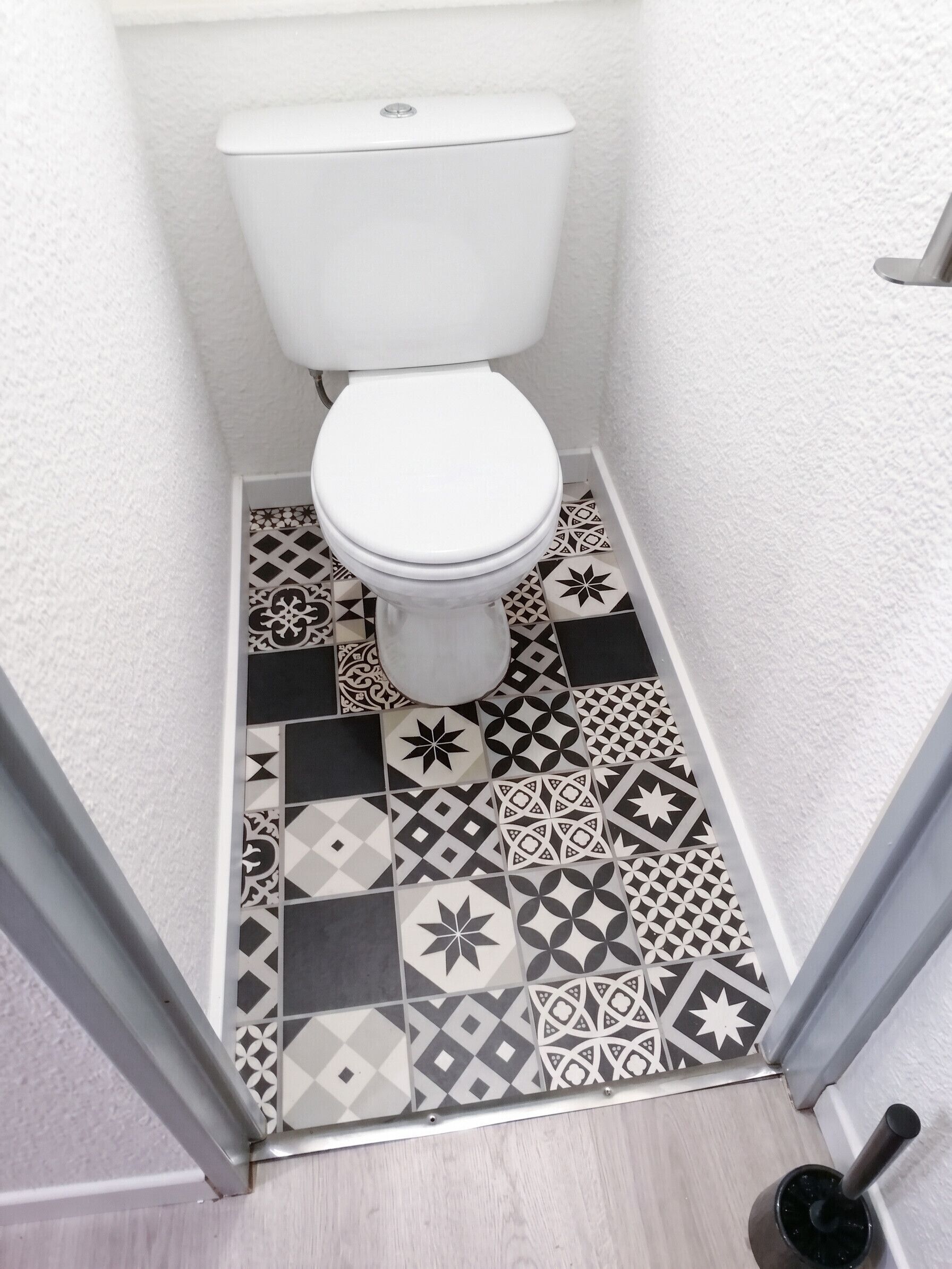 A small bathroom with a toilet, patterned floor tiles, and a wooden floor.