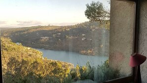 A bedroom with a large window overlooking a valley and a lake. There is a bed with a white quilt and pillows, and a pink lamp on a bedside table. - Architect's house, rustic style, will seduce you by its environment  (Esparron-De-Verdon)