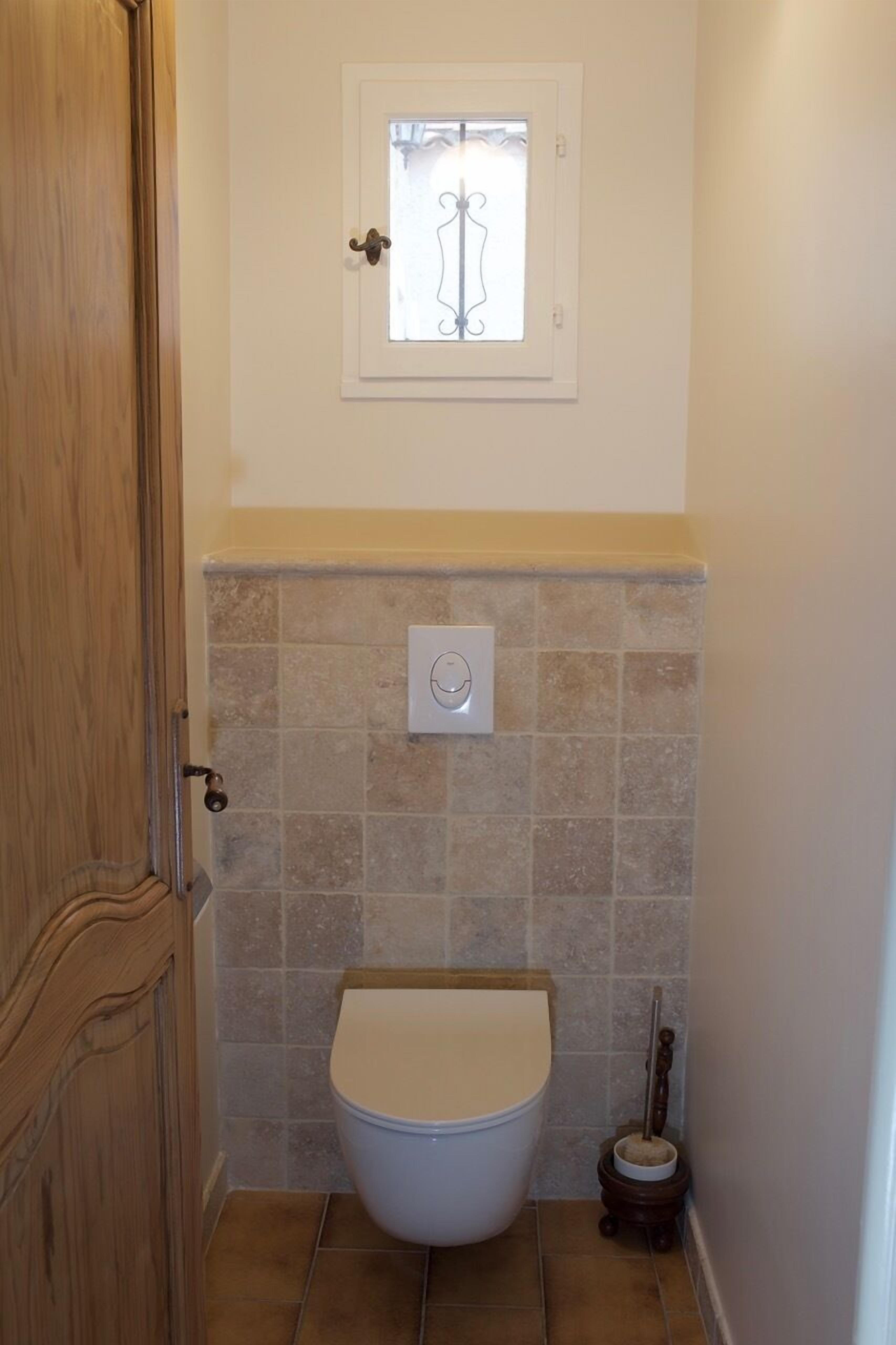 Une petite salle de bain comprenant des toilettes, une fenêtre ornée d’une grille décorative et un mécanisme de chasse d’eau fixé au mur.