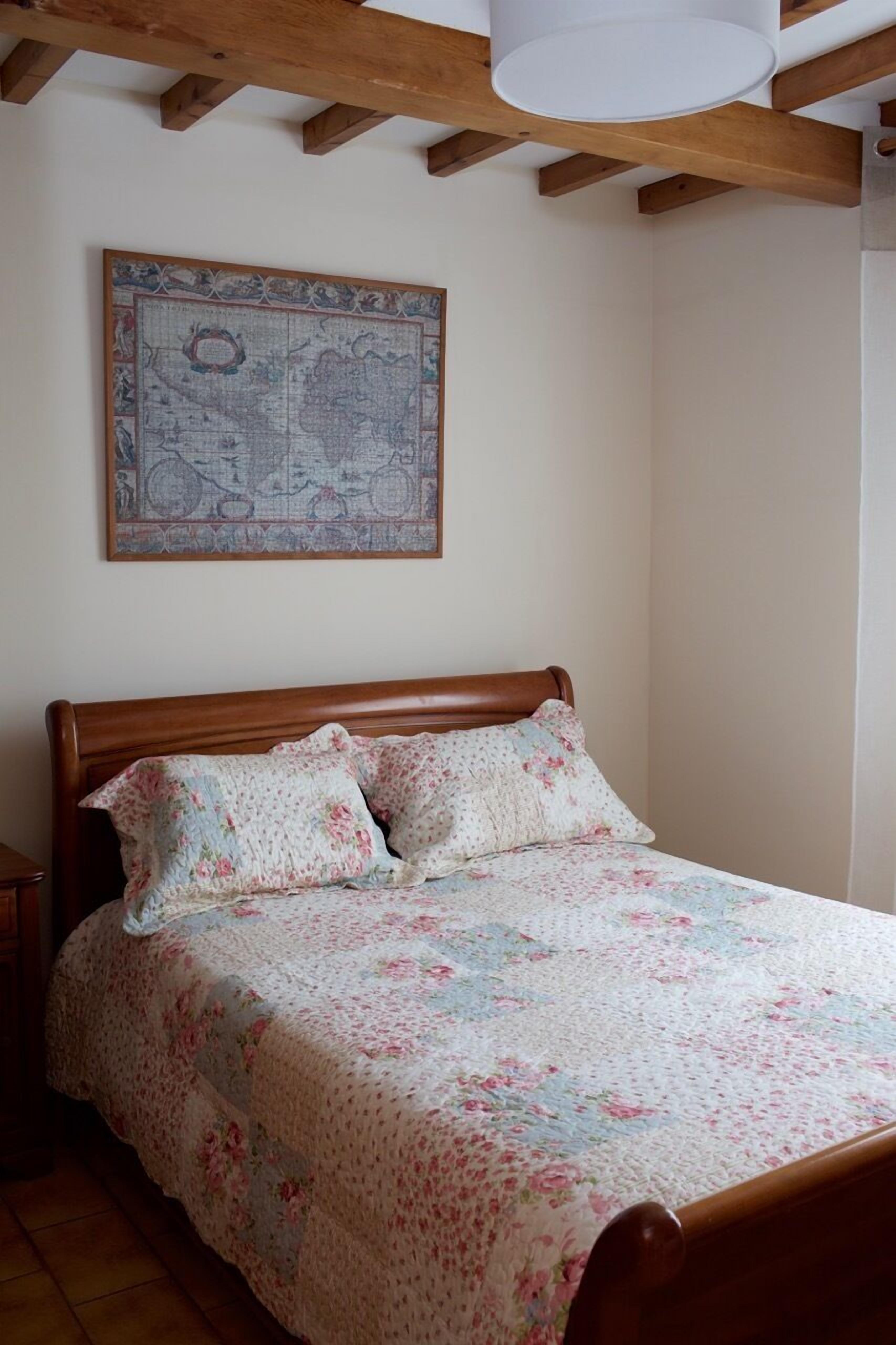 A bedroom with a wooden bed, floral bedding, a map on the wall, and a wooden ceiling beam.