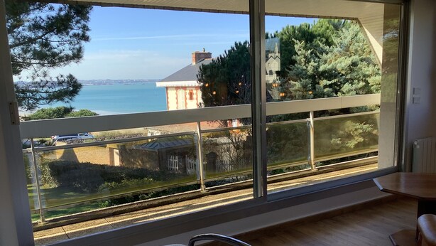 A room with a large window overlooking a scenic view of a body of water, a train track, and a distant building. - Beautiful family home with sea view and beach access (Lancieux)
