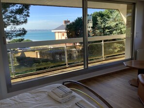 A room with a large window overlooking a scenic view of a body of water, a train track, and a distant building. - Beautiful family home with sea view and beach access (Lancieux)