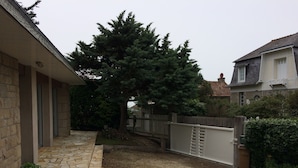 A stone-paved patio with a wooden fence, a large tree, and a house with a tiled roof. - Beautiful family home with sea view and beach access (Lancieux)