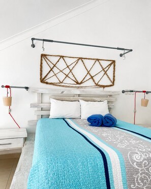Ein ordentlich eingerichtetes Schlafzimmer mit Bett, Kissen, Kopfteil, einer Wandleuchte und einem Wandbild.