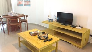 Un salon avec une table en bois, des chaises, un téléviseur posé sur un meuble en bois, et une petite étagère sur laquelle se trouvent une bouteille et une télécommande.