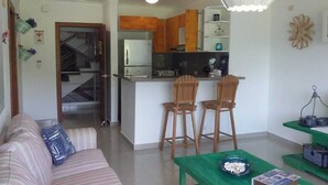 Un salon avec un canapé, une table verte et deux chaises en bois. On aperçoit à l’arrière-plan un coin cuisine avec un réfrigérateur, une cuisinière et des placards en bois.