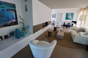 A modern living room with a white sofa, a white armchair, a coffee table, and a fireplace. There is a painting on the wall and a plant on the side.