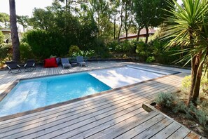 A wooden deck with a swimming pool, lounge chairs, and a red cushion.