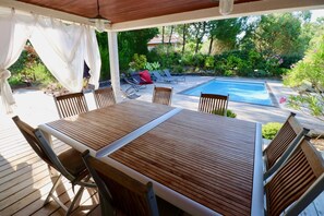 A wooden deck with a table and chairs, overlooking a swimming pool and lush garden.