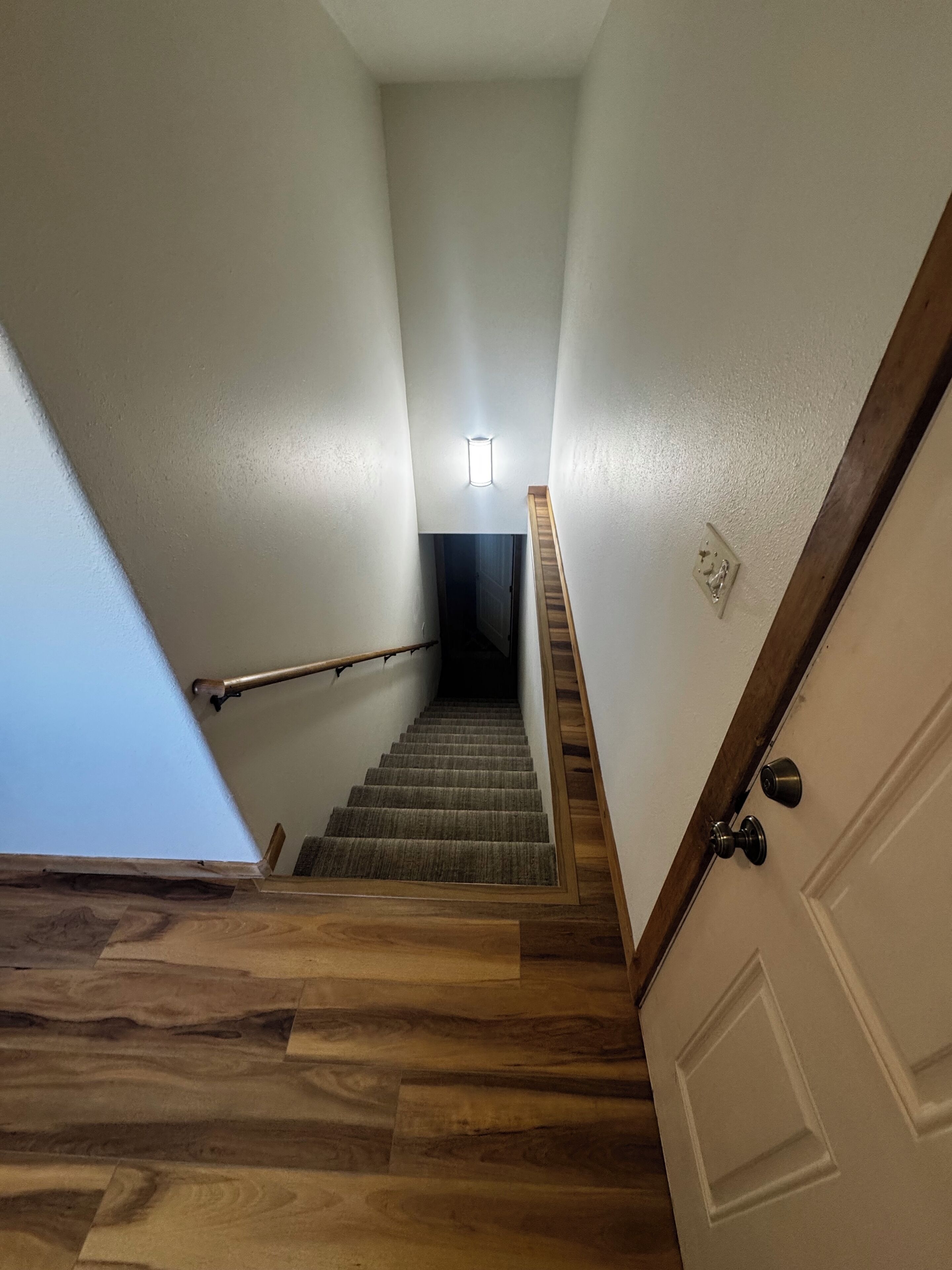 Een trap met houten treden en een leuning, die leidt naar een deur. Op de muur is een lichtschakelaar en een rookmelder te zien.
