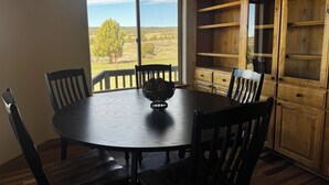 A dining area with a round table, chairs, a wooden cabinet, and a view of a green landscape through a window. - Gateway to Yellowstone, Island Park  (Ashton)