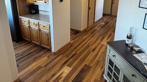 A hallway with wooden flooring, a kitchen area with wooden cabinets, and a dining area with a cabinet and a bottle on the counter. - Gateway to Yellowstone, Island Park  (Ashton)
