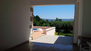 An interior space leading to a balcony with a pool, surrounded by greenery and mountains in the background.