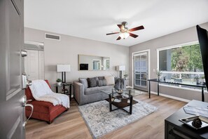 A living room with a grey sofa, a red chair, a coffee table, a ceiling fan, and a view through large windows. - A Perfect Retreat (Chandler)