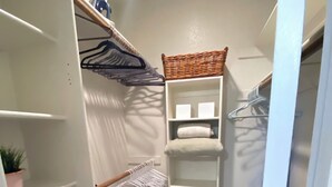 A narrow hallway with a white closet, shelves, a wicker basket, a wicker basket on a shelf, a white shelf, a white shelf with a white cushion, a wicker basket on the floor, a white trash bin, and a door.