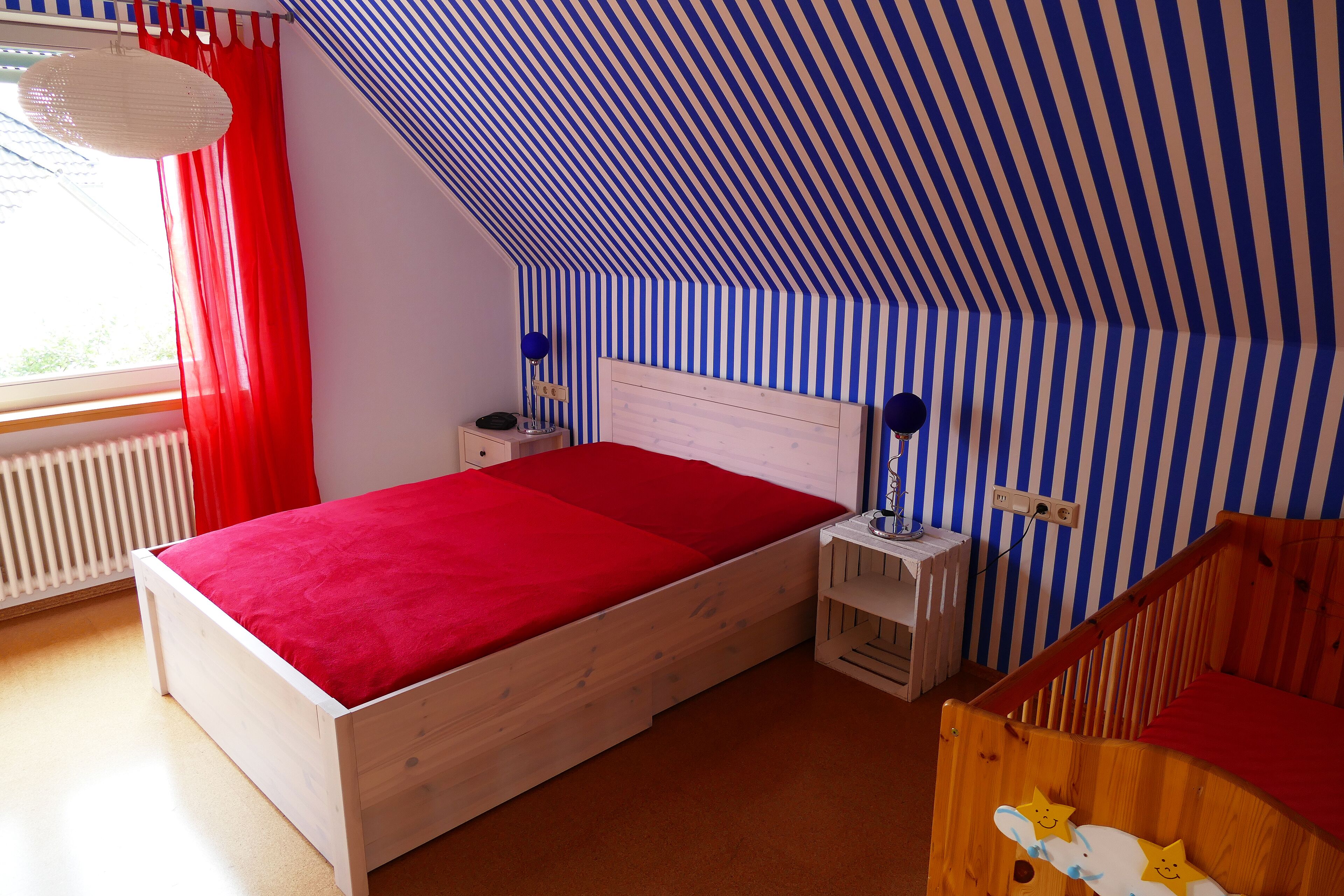 Un dormitorio con cama de madera, ropa de cama roja, techo a rayas y una ventana con cortinas rojas.
