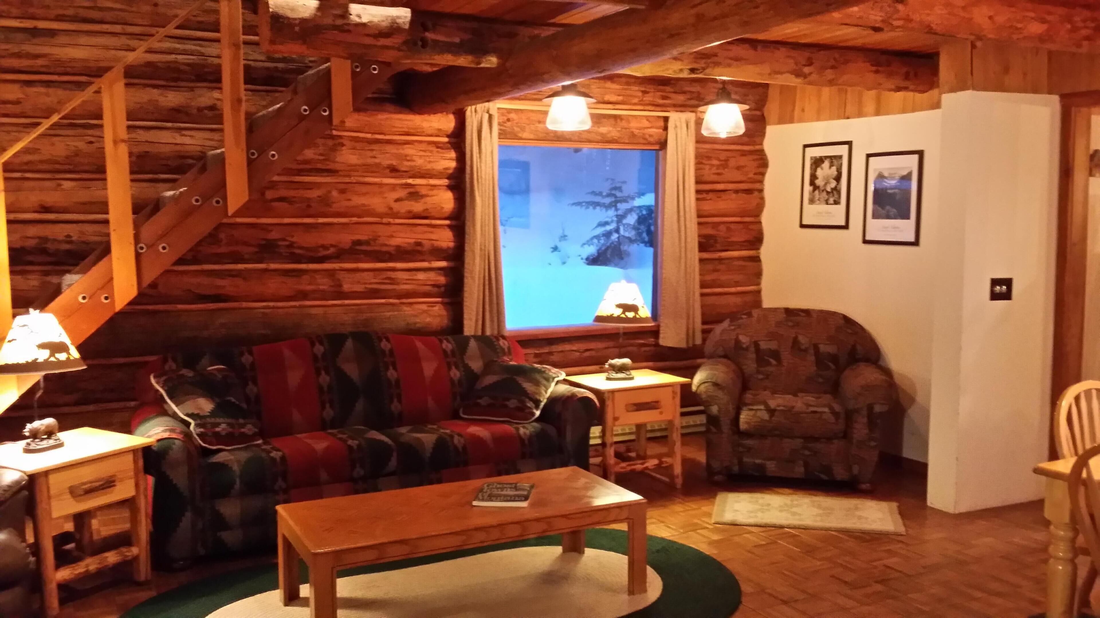 Interior acogedor de una cabaña de troncos con una escalera de madera, un sofá, un sillón estampado, una mesita y una ventana con vistas a un paisaje nevado.