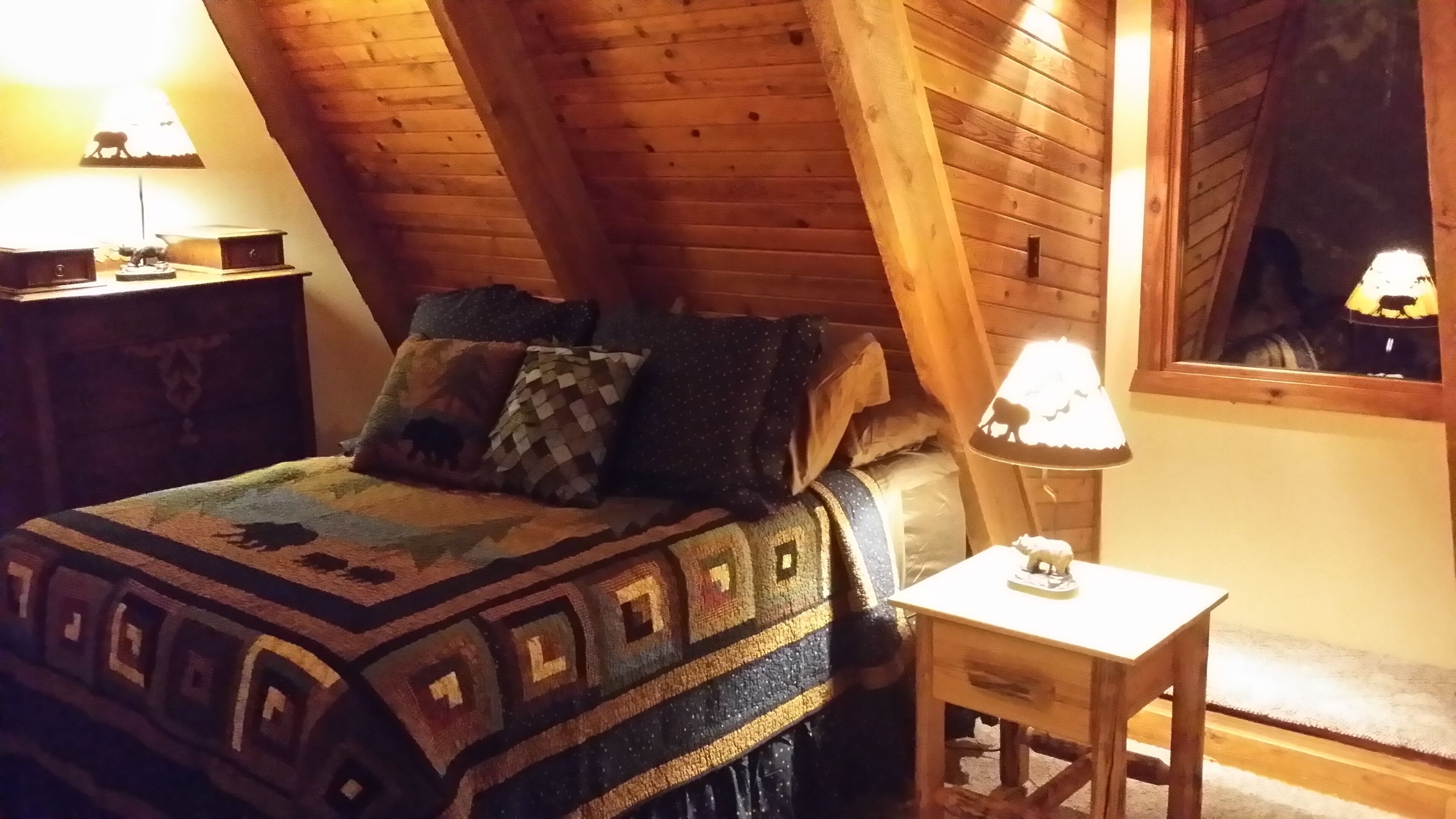 Un dormitorio acogedor en el ático, con techo de madera, una cama con ropa de cama estampada, una mesita de noche con lámpara y una pequeña mesa.