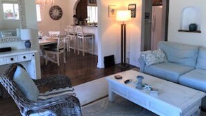 A living room with a white sofa, a coffee table, and a wicker chair. There is a dining area with chairs and a table in the background.