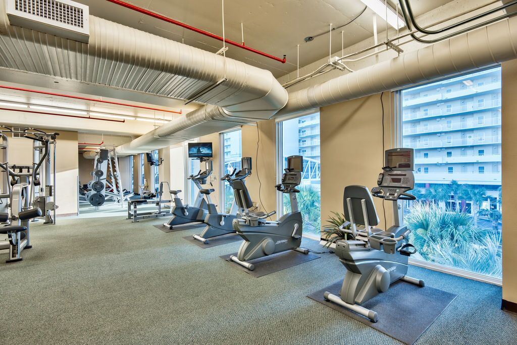 A gym with cardio machines, weightlifting equipment, and large windows offering a view of a building and greenery.
