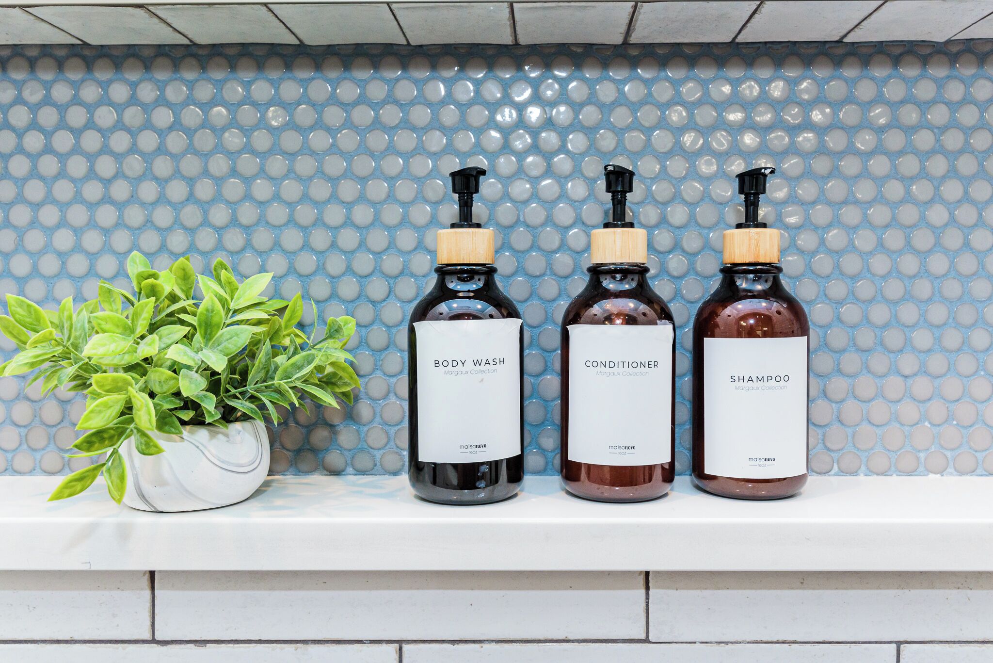 Trois bouteilles de gel douche, d’après-shampoing et de shampoing sont posées sur une étagère blanche, devant un mur recouvert de carreaux bleus.
