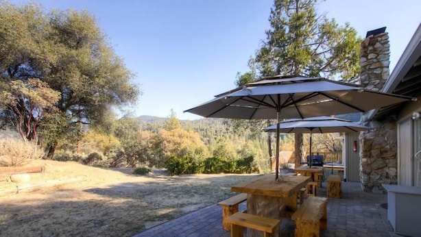 An outdoor patio with wooden tables and benches, a large umbrella, and a stone chimney. - Lazy Bear Lodge (Oakhurst)