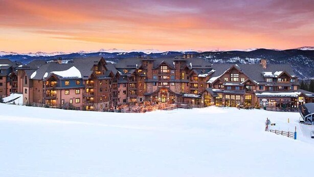 A large lodge with multiple buildings, a snow-covered landscape, and mountains in the background.